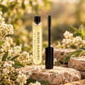 Eyelash + Eyebrow Growth Serum bottle with its black wand applicator displayed beside it among white flowers.