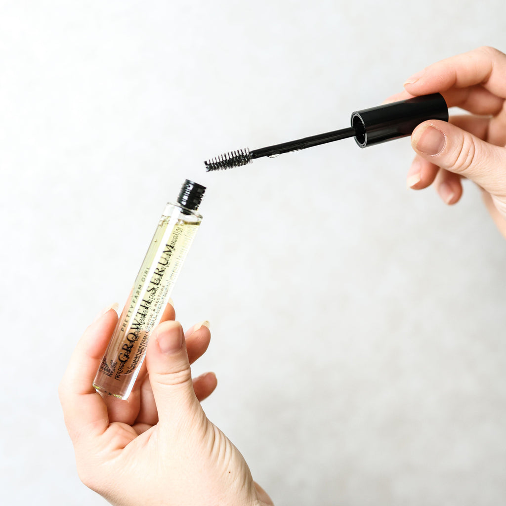 A hand holds the Eyelash + Eyebrow Growth Serum tube beside its black spoolie applicator.